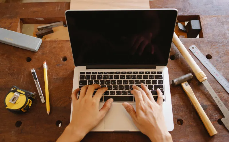 Hands typing on a laptop at a woodworker's bench, with a tape measure, pencil and hand tools nearby